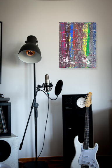Schreibtisch mit Mikrofon, Gitarre, Lampe und farbenfrohem Kunstwerk an der Wand.